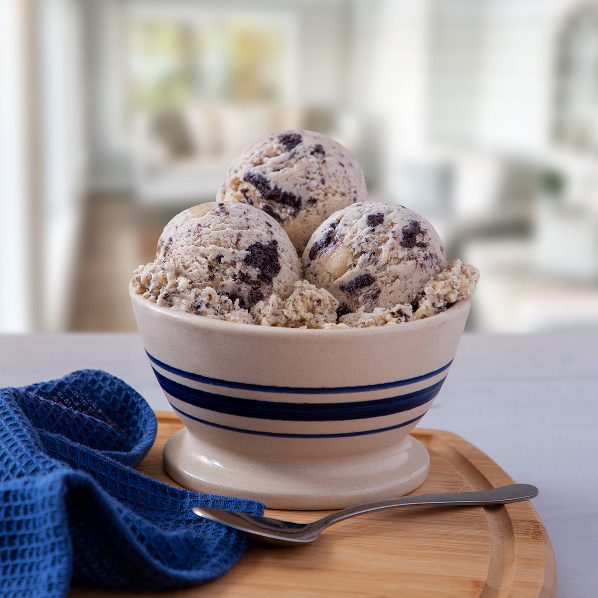 Blue Bell Cookie Two Step Ice Cream in a white ceramic bowl with blue stripes, set on a wooden board with a spoon and blue cloth napkin in a bright kitchen setting.