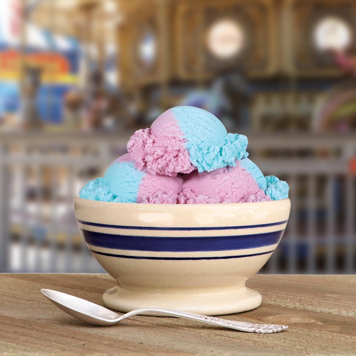 Bowl of Blue Bell Cotton Candy Ice Cream in pink and blue with a spoon in a carnival setting.