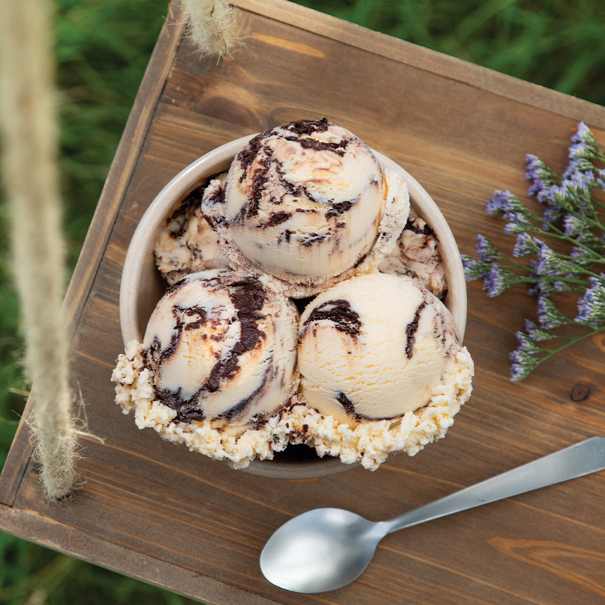 Bowl of Blue Bell Homemade in the Shade Ice Cream in a bowl on a wooden tray, with a spoon and purple flowers nearby.