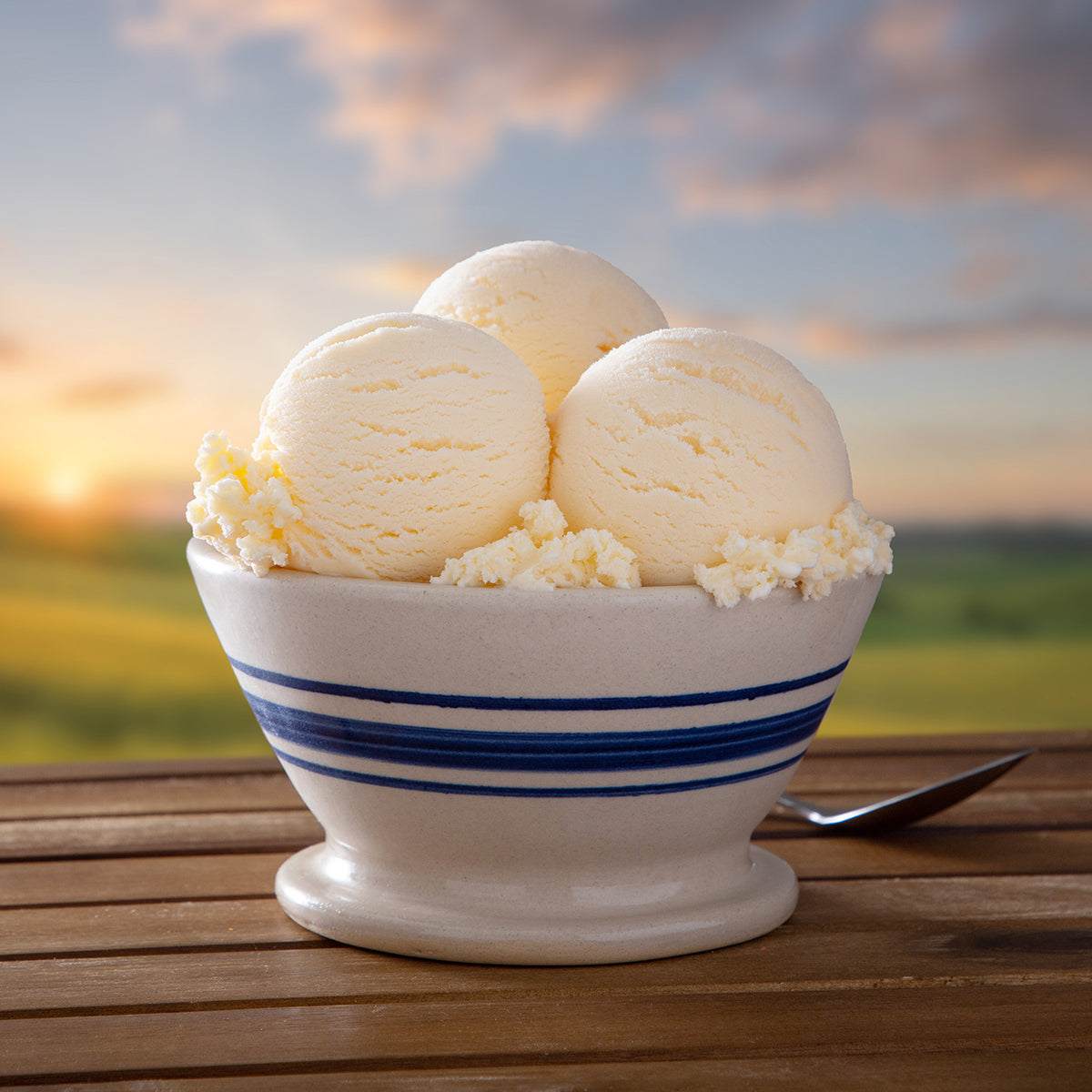 Bowl of Blue Bell Homemade Vanilla Ice Cream on a wooden table with a spoon and a morning sunrise in the background.