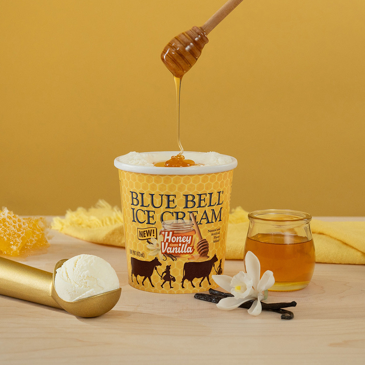 Blue Bell Honey Vanilla Ice Cream carton with honey drizzle, honeycomb, vanilla beans and a golden scoop of ice cream on a gold background.