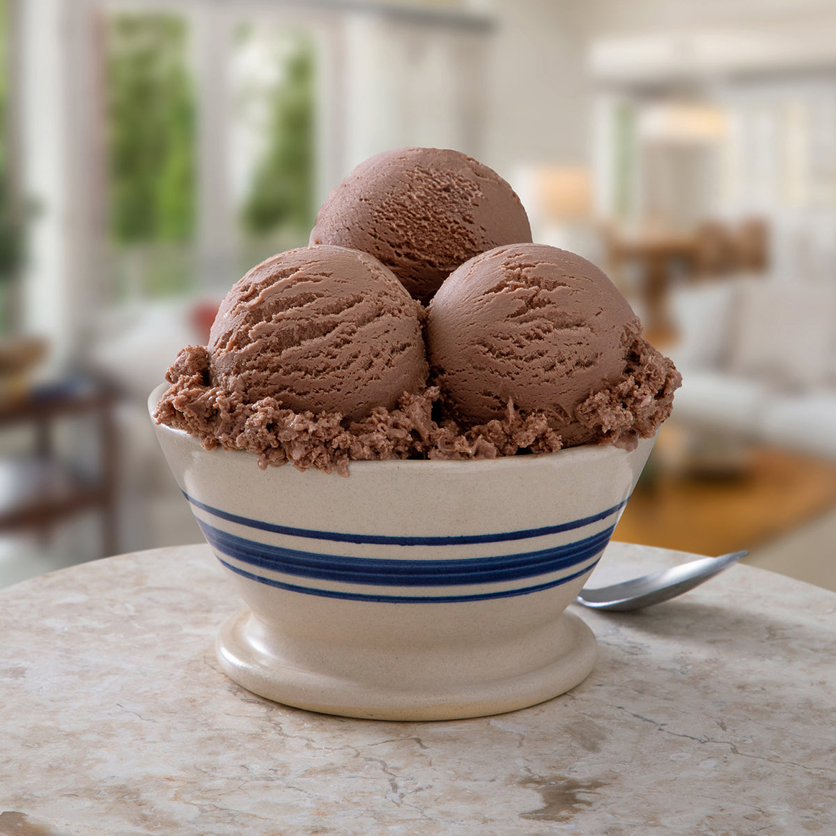 Blue Bell Milk Chocolate Ice Cream in a white ceramic bowl with blue stripes, filled with chocolate crumble, on a marble table with a spoon in a blurred indoor setting.