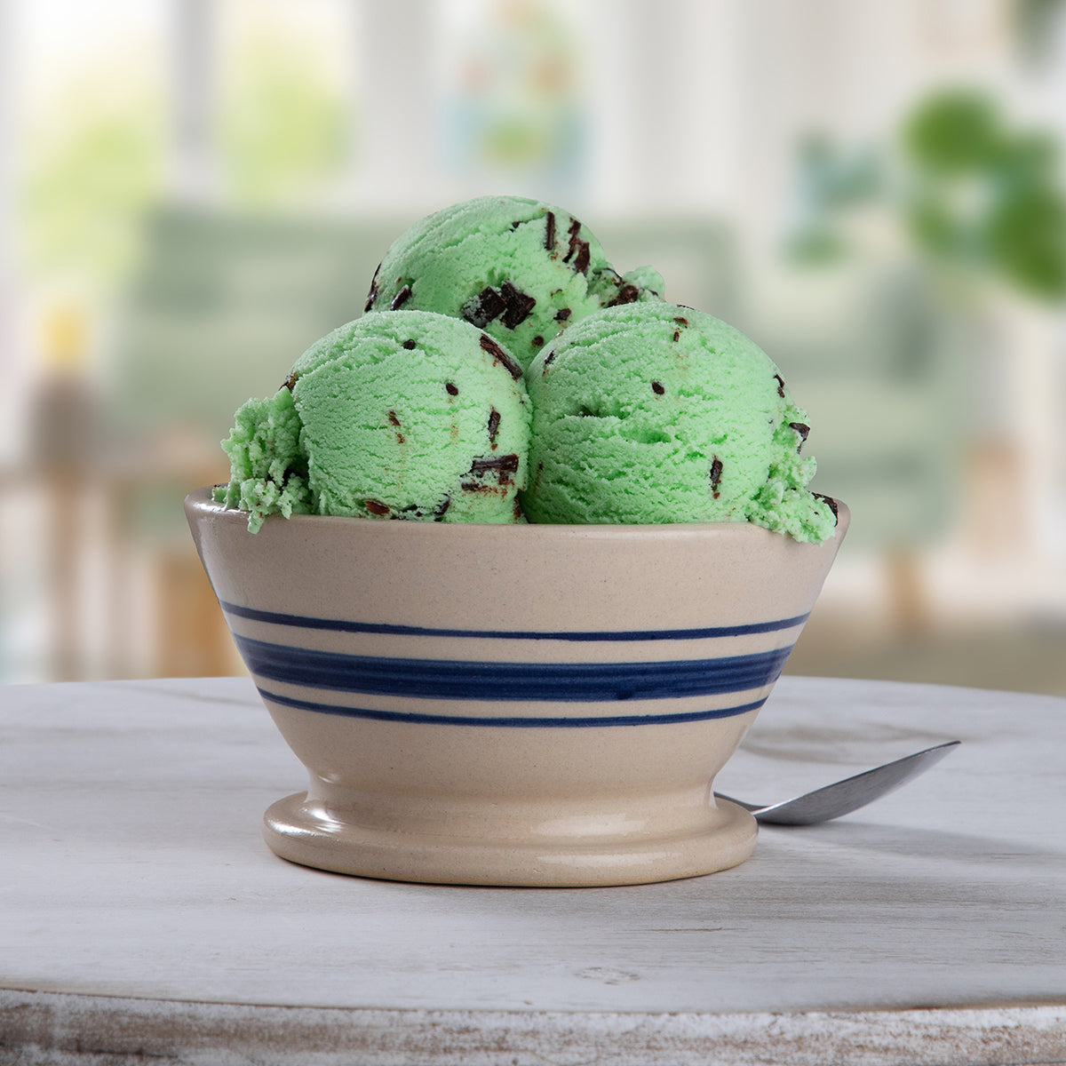 Bowl of Blue Bell Mint Chocolate Chip Ice Cream with a spoon on a gray wooden table and a blurred background setting.
