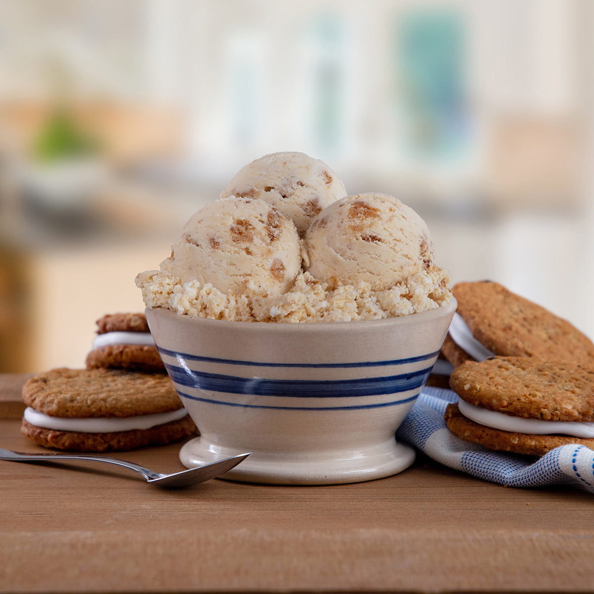 Blue Bell Oatmeal Cream Pie Ice Cream with crumbled cookie pieces in a small white bowl with blue stripes, styled on a wooden surface with oatmeal cream pie cookies and a spoon.