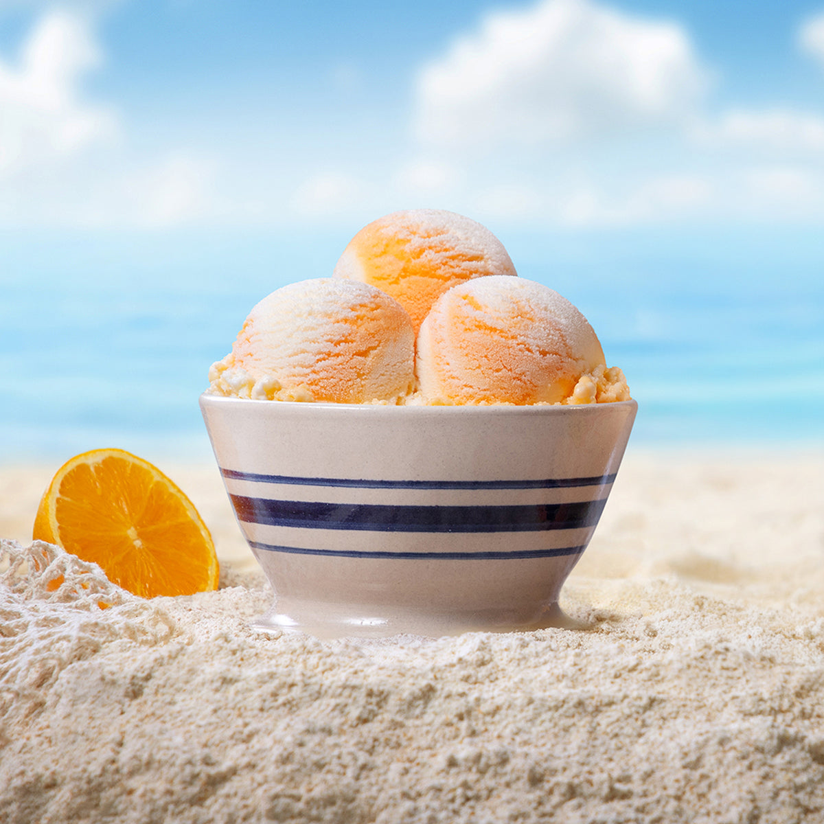 Bowl of Blue Bell Orange Swirl Ice Cream on a beach with sand, oranges and a blue sky.