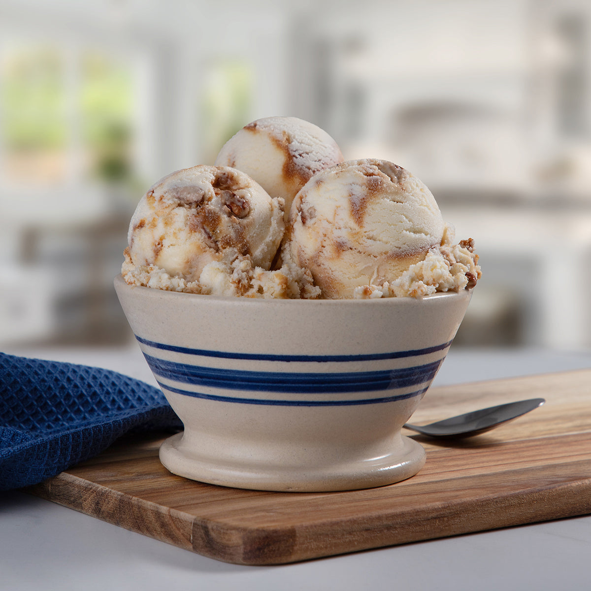 A bowl of Blue Bell Pecan Pralines n Cream Ice Cream on a wooden cutting board with a blue towel and spoon.  