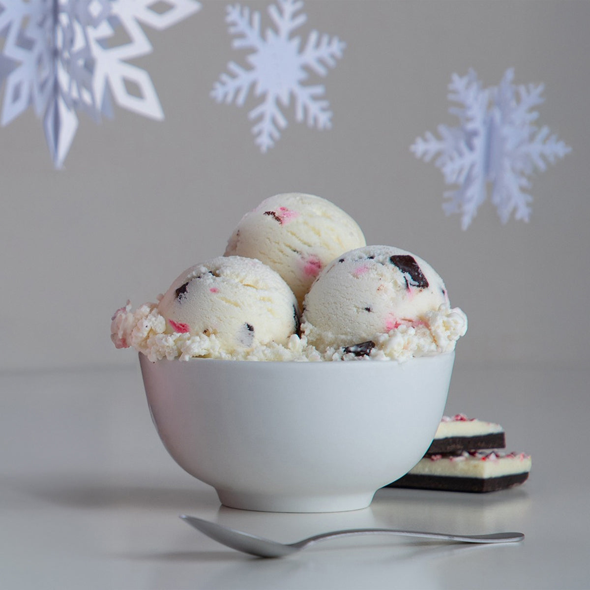 White bowl of Blue Bell Peppermint Bark Ice Cream with peppermint bar and a spoon on a white background with snowflakes.