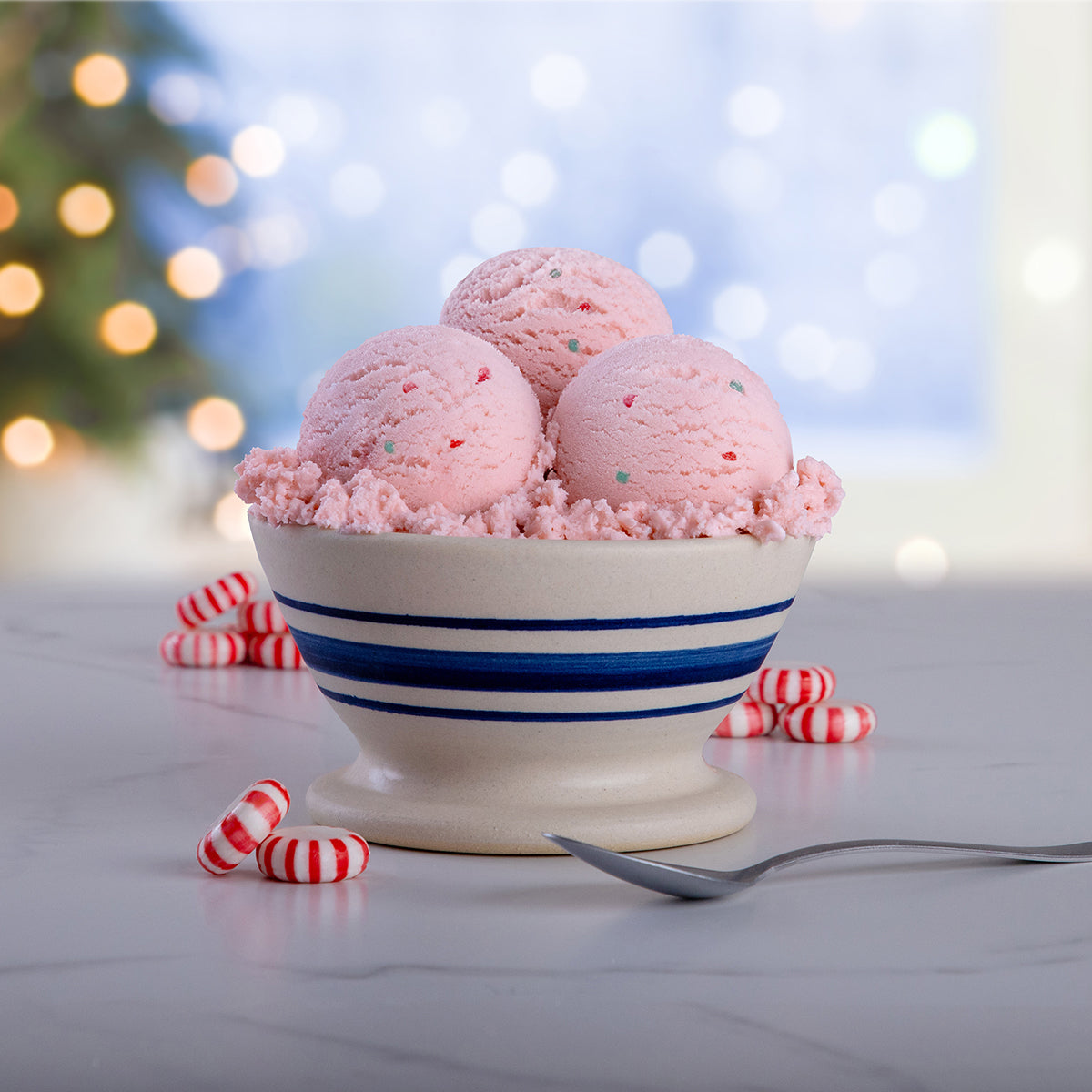 Bowl of Blue Bell Peppermint Ice Cream on a marble table with a spoon, red and white peppermints and a Christmas background.