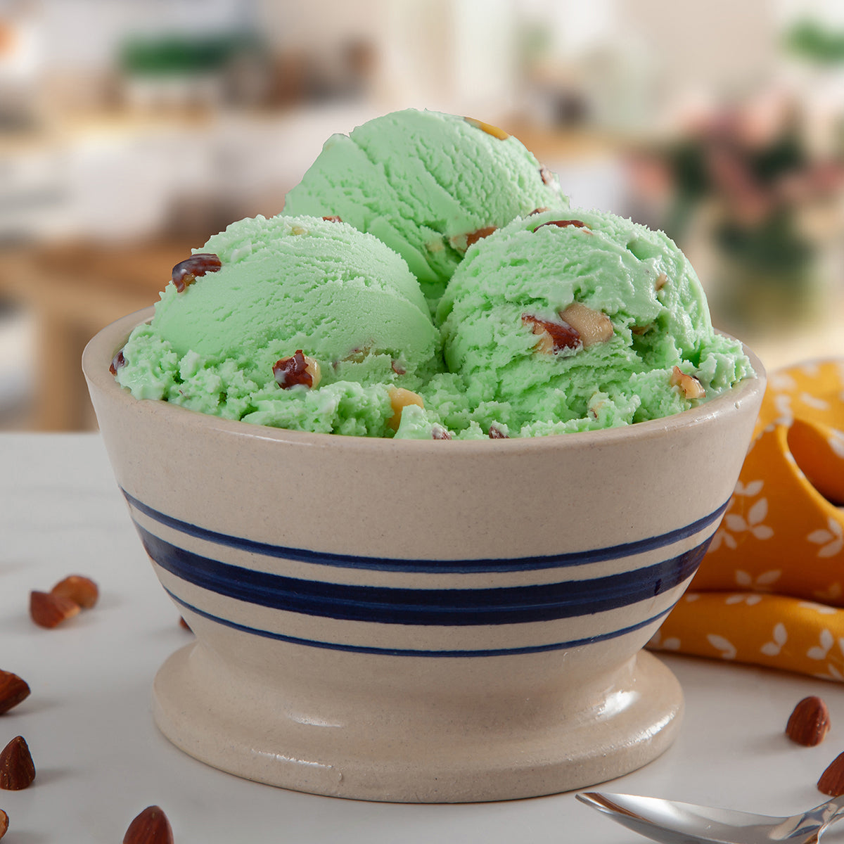 Bowl of Blue Bell Pistachio Almond Ice Cream with roasted almonds, spoon, golden towel in a blurred kitchen background.