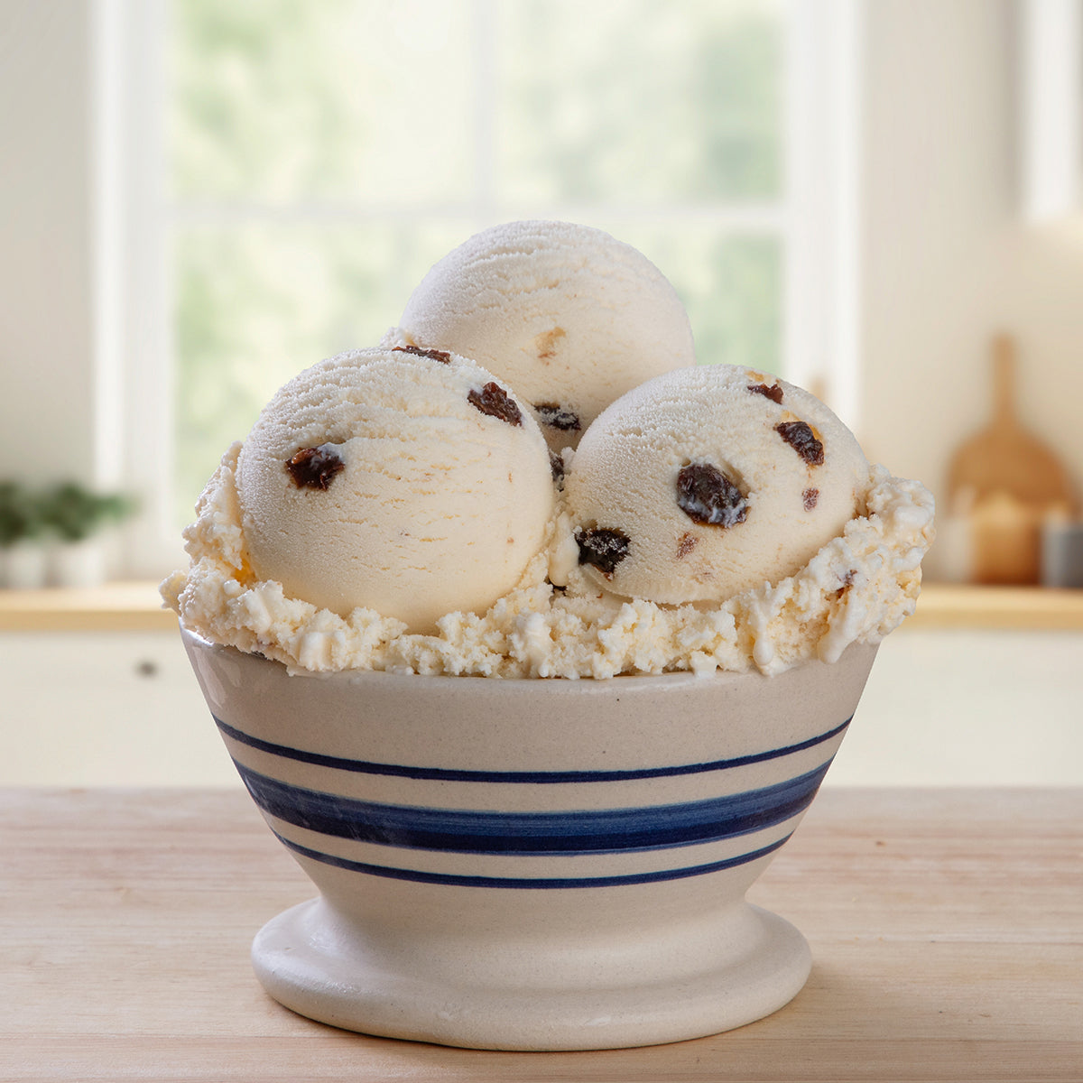 Blue Bell Rum Raisin Ice Cream served in a white ceramic bowl with blue stripes, set on a wooden table with a softly blurred kitchen background.