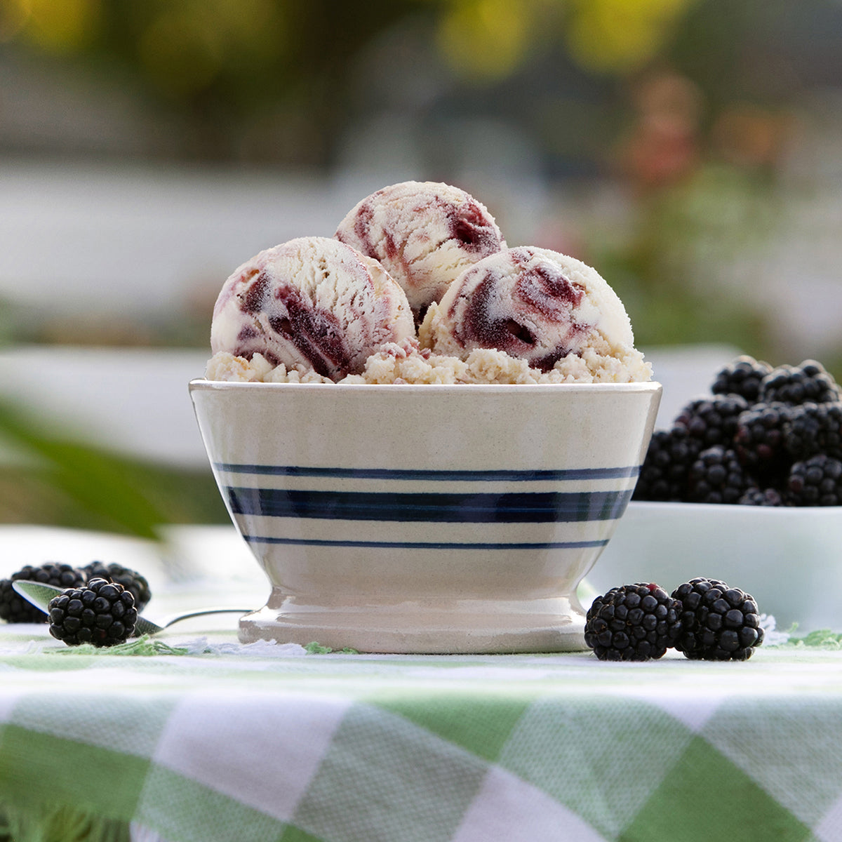 A bowl of Blue Bell Southern Blackberry Cobbler ice cream with blackberry swirls, served on a green and white checkered tablecloth with fresh blackberries around it.