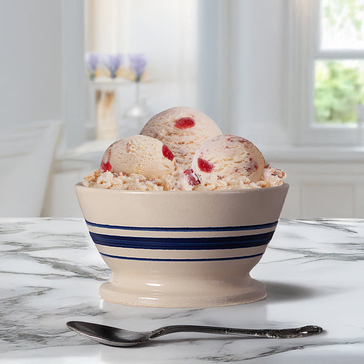 Bowl of Blue Bell Strawberries and Homemade Vanilla Ice Cream with a spoon on a marble countertop in a blurred kitchen setting. 