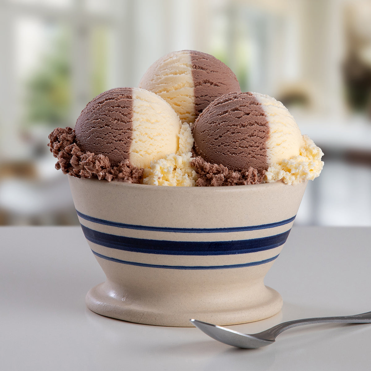 A bowl of Blue Bell The Great Divide Ice Cream featuring chocolate and vanilla ice cream with a spoon on a light tabletop against a blurred indoor background.