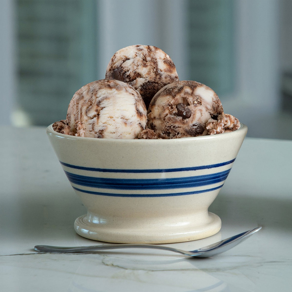 Three scoops of Blue Bell Tin Roof Ice Cream in a cream-colored ceramic bowl with blue stripes, set on a light marble surface with a silver spoon in front.