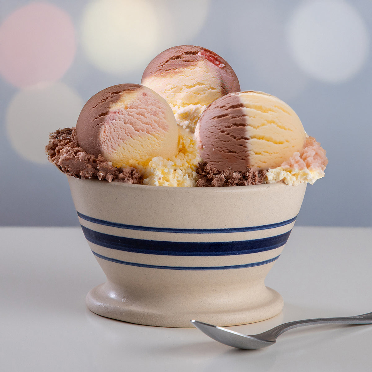 Three scoop of Blue Bell Ultimate Neapolitan Ice Cream in a cream-colored ceramic bowl with blue stripes. A metal spoon rests in the foreground against a softly blurred pastel bokeh background.