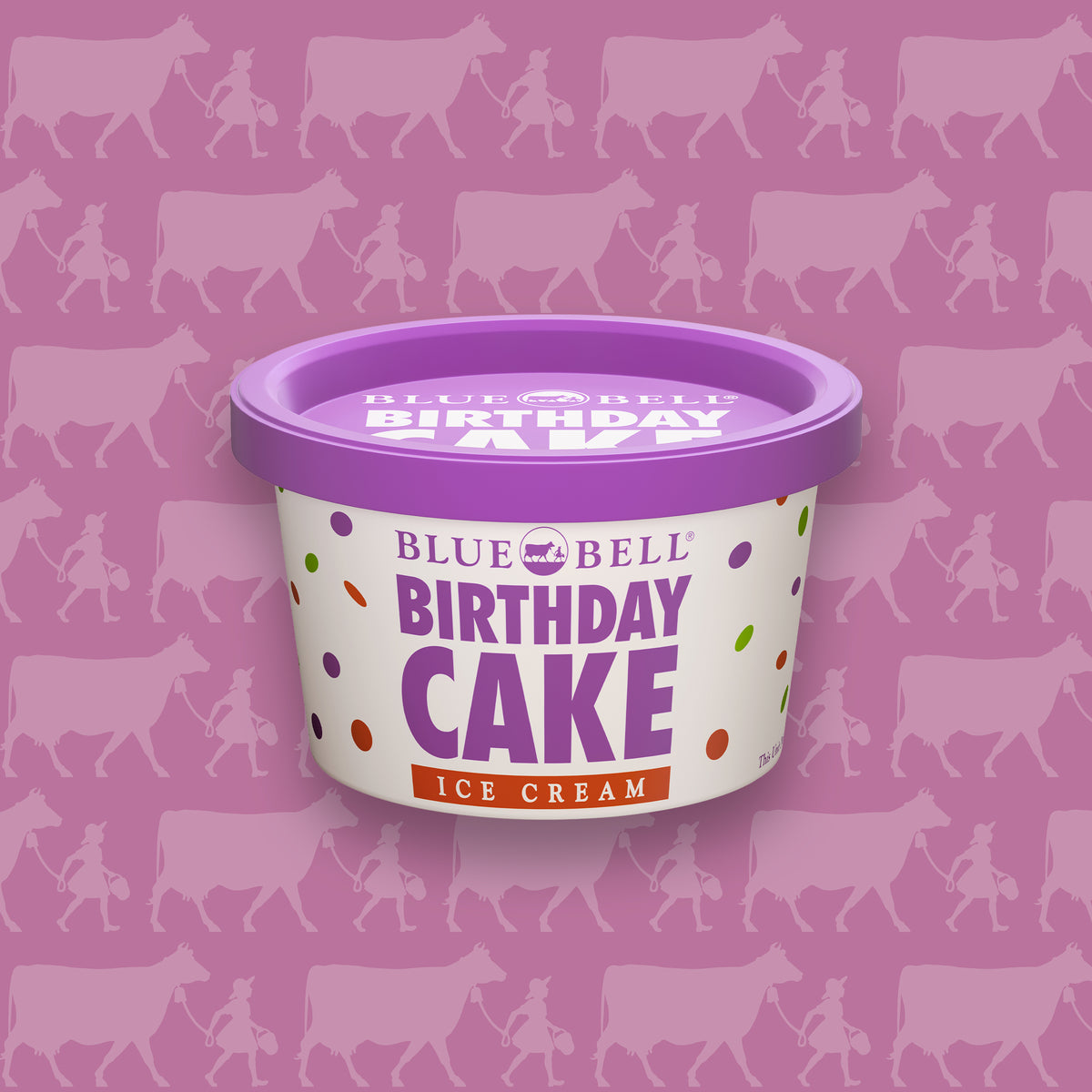 A cup of Blue Bell Birthday Cake Ice Cream centered on a pink background patterned with cow silhouettes. The cup is white with purple, red, and multicolor polka dots, and the product name is printed prominently on the front.