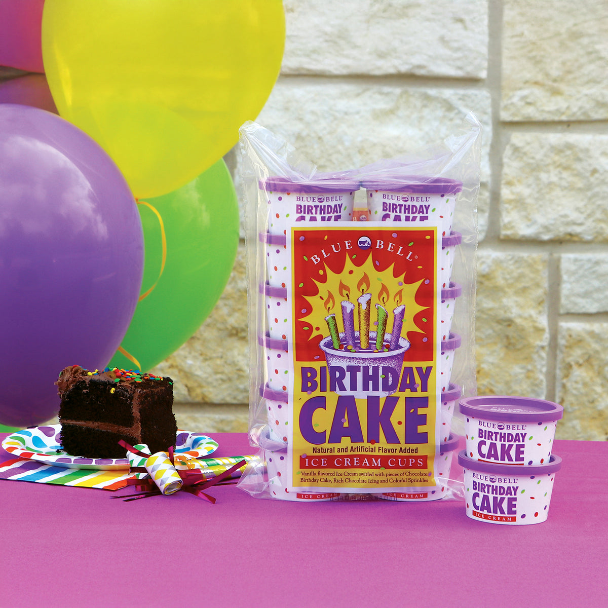 A packaged set of Blue Bell 'Birthday Cake' ice cream cups displayed on a pink table with balloons, a slice of chocolate cake, and a party hat prop. The main product is a clear plastic bag containing multiple cups with purple lids and a colorful front label, with two smaller cups shown in front.