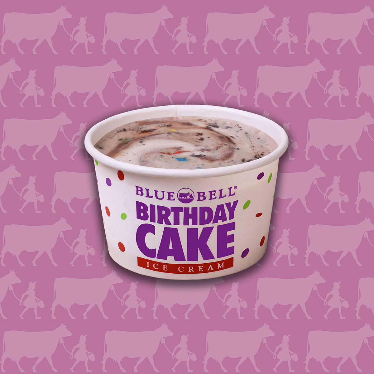 A cup of Blue Bell Birthday Cake Ice Cream centered on a pink background patterned with cow silhouettes. The cup is white with purple, red, and multicolor polka dots, and the product name is printed prominently on the front.