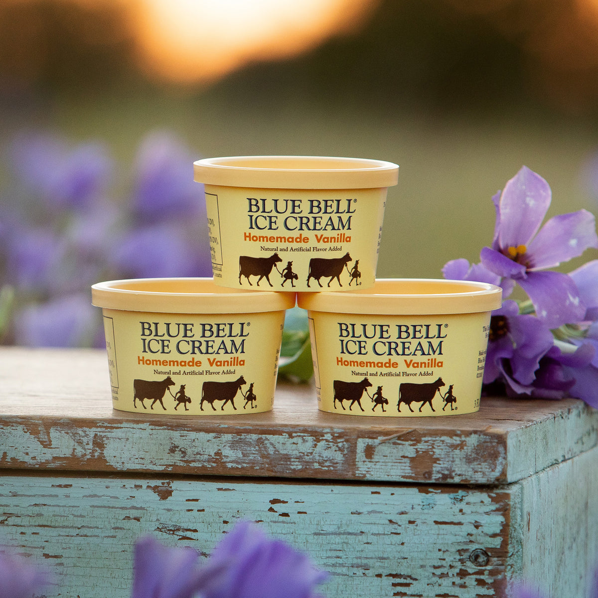 Three Blue Bell Ice Cream Homemade Vanilla cup containers arranged in a pyramid on a rustic distressed wooden surface, with purple bluebell flowers in the foreground and a softly blurred outdoor background.