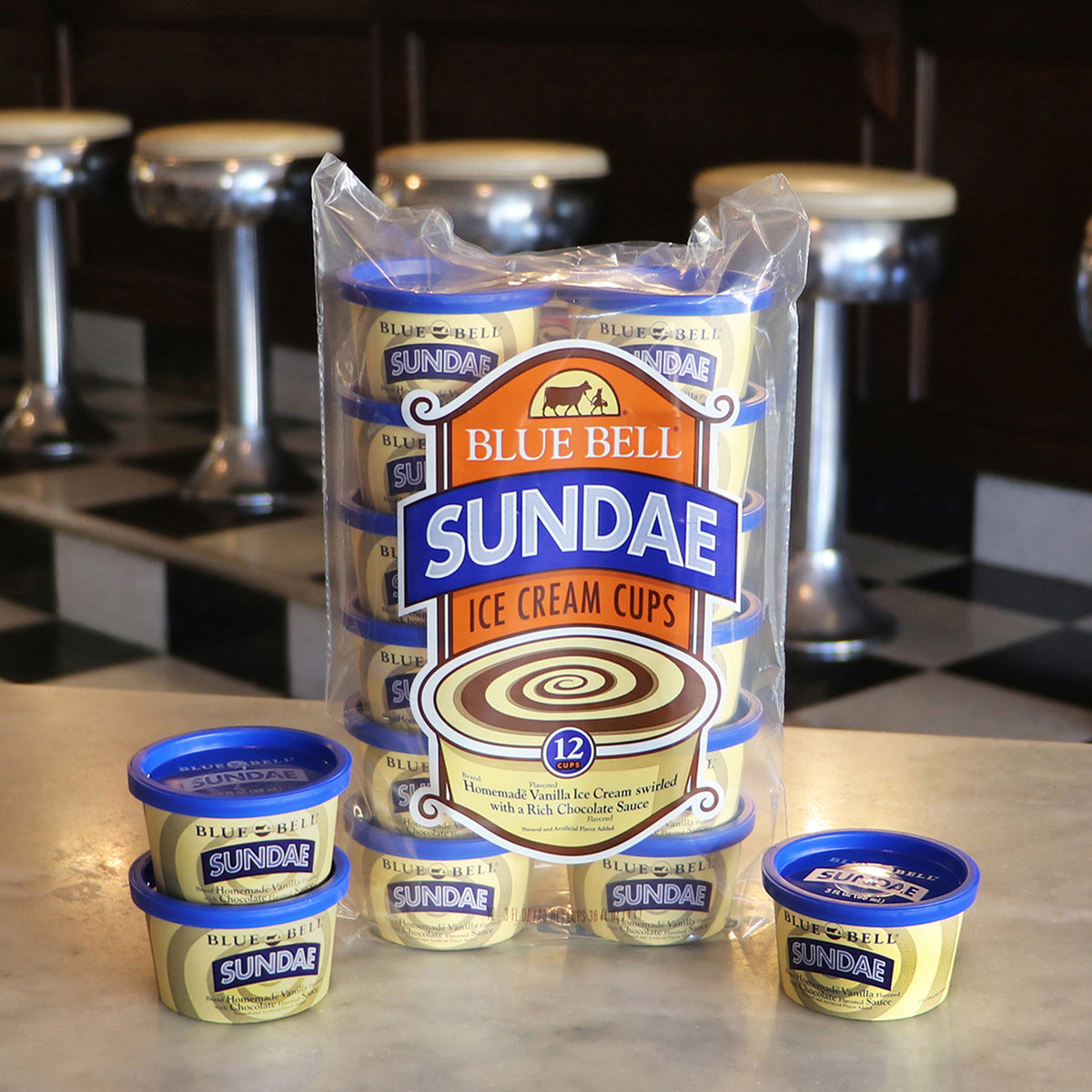 A 12-pack of Blue Bell Sundae Ice Cream Cups in a clear plastic bag, displayed on a counter with four individual cups in front and blurred bar stools in the background.