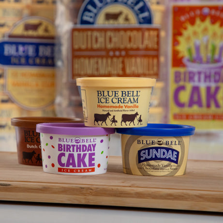 Collection of Blue Bell ice cream cups on a wooden surface with various flavors displayed.