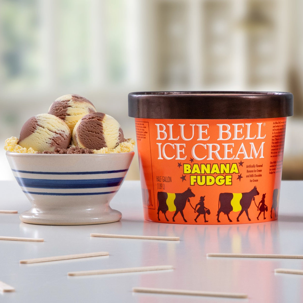 A tub of Blue Bell Ice Cream Banana Fudge next to a small bowl of vanilla ice cream with chocolate swirls on a tabletop.