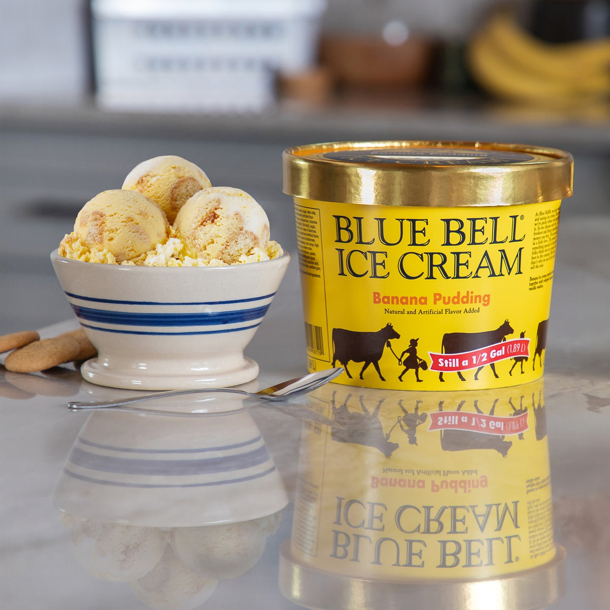 A tub of Blue Bell Ice Cream in Banana Pudding flavor stands on a reflective surface next to a small bowl of banana pudding ice cream with banana pieces, vanilla wafers, and a spoon in a kitchen setting.