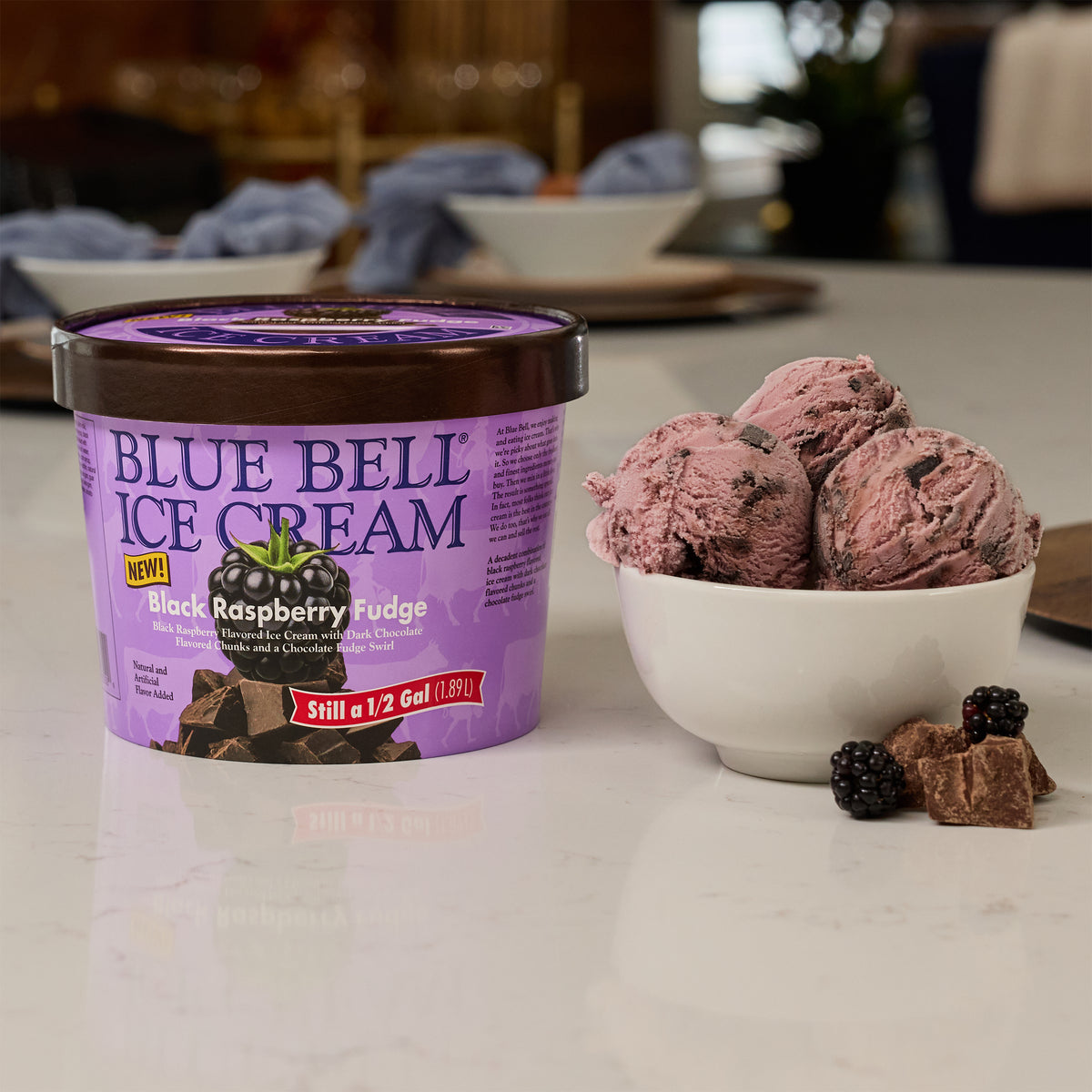 A tub of Blue Bell Ice Cream in Black Raspberry Fudge flavor sits on a white marble countertop next to a white bowl filled with scoops of the dark berry ice cream, with a few blackberries and chocolate pieces nearby.