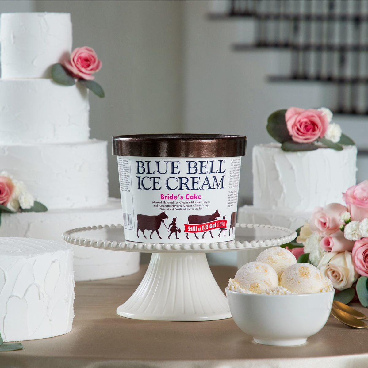Half Gallon of Blue Bell Bride's Cake in a white carton on a white cake pedestal with a bowl of ice cream, flowers and wedding cakes in the background. 
