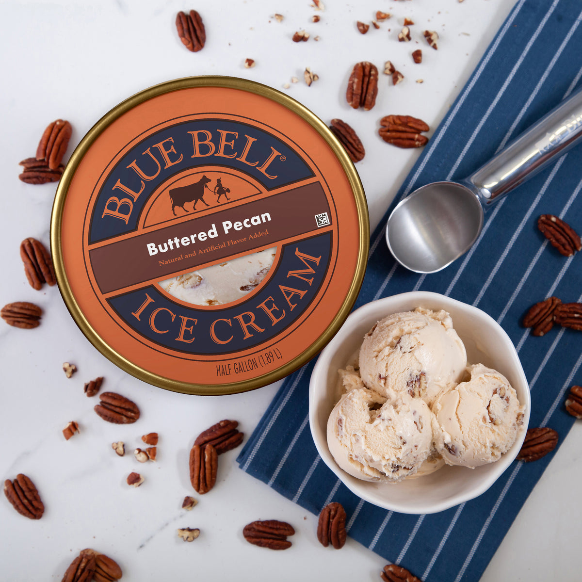 Blue Bell Buttered Pecan Ice Cream in a round tub, styled on a white marble surface with scattered pecans, a white bowl of scoops, a blue striped napkin, and a metal ice cream scoop.
