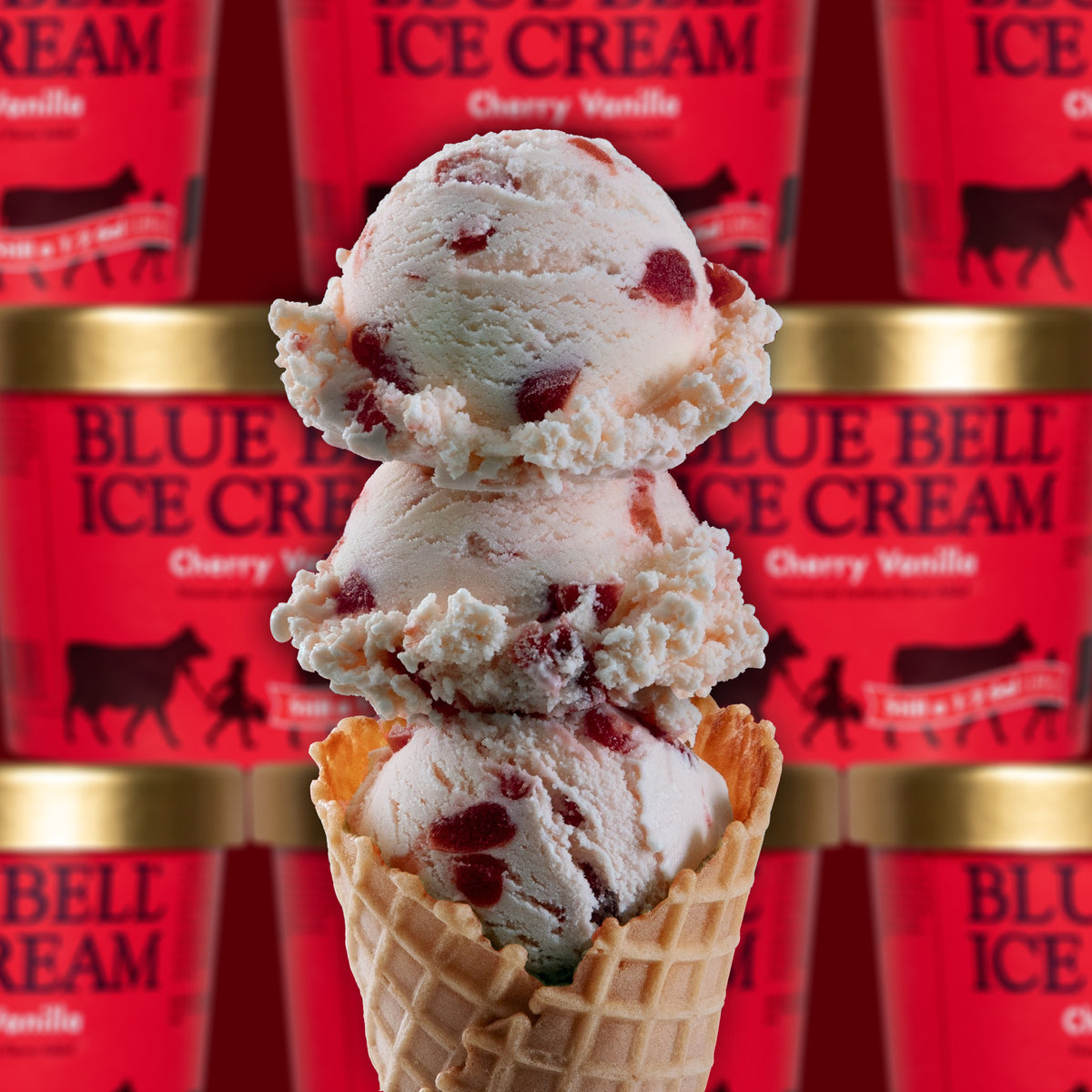 A close-up of a waffle cone filled with scoops of Blue Bell Ice Cream in Cherry Vanilla flavor, with multiple red Blue Bell Ice Cream tubs visible in the blurred background.