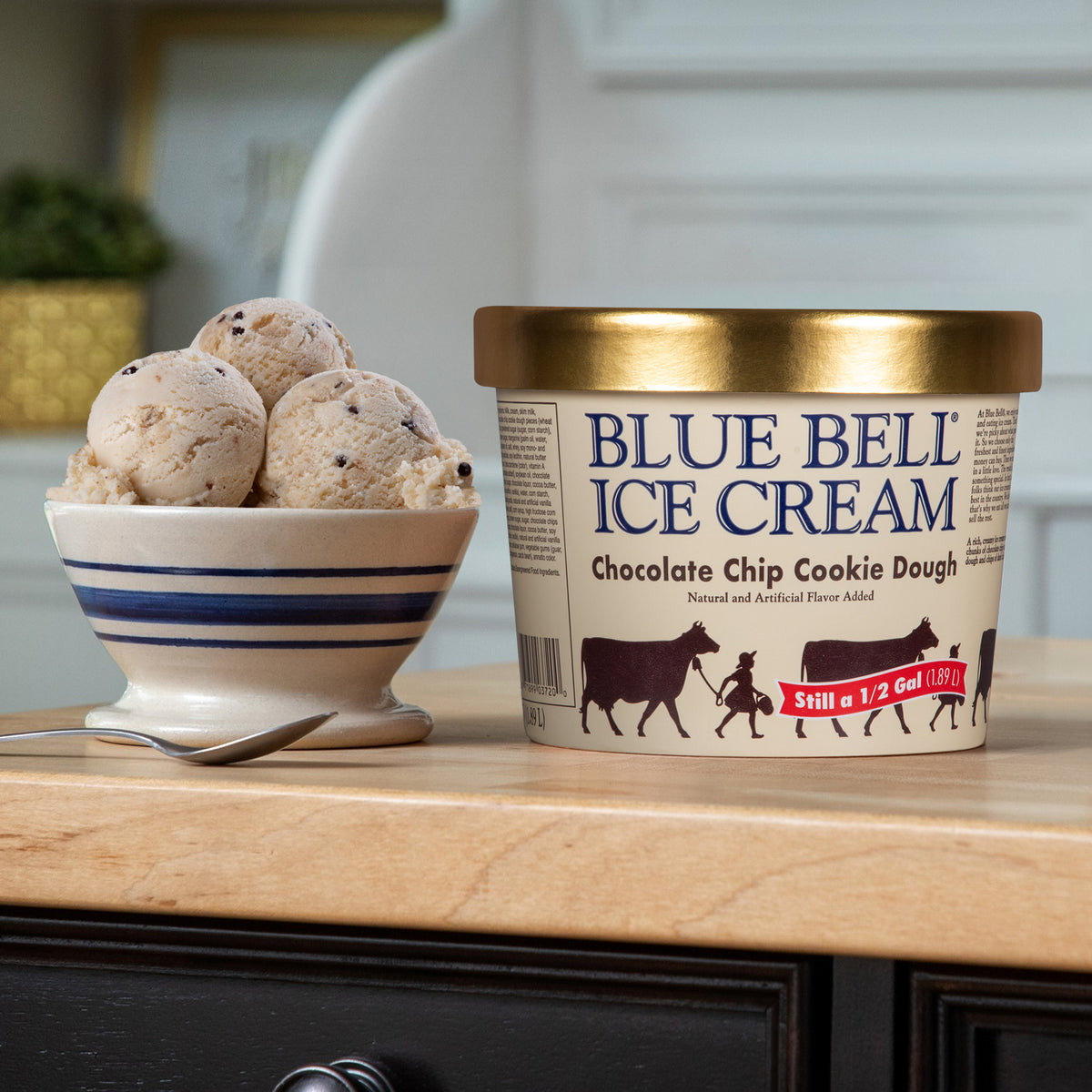 A tub of Blue Bell Ice Cream Chocolate Chip Cookie Dough stands on a wooden counter next to a bowl of scoops of ice cream in a kitchen setting.