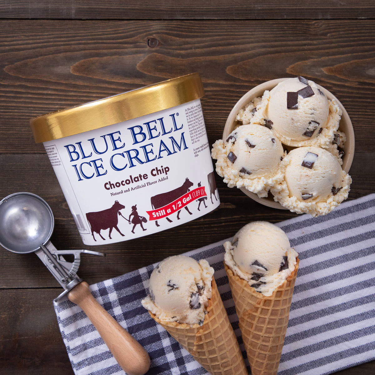 A tub of Blue Bell Ice Cream Chocolate Chip flavor is arranged on a dark wood surface with two waffle cones, a bowl of scoops, and an ice cream scoop. The Blue Bell Ice Cream container is the main product, with the ice cream itself shown in cones and a bowl.