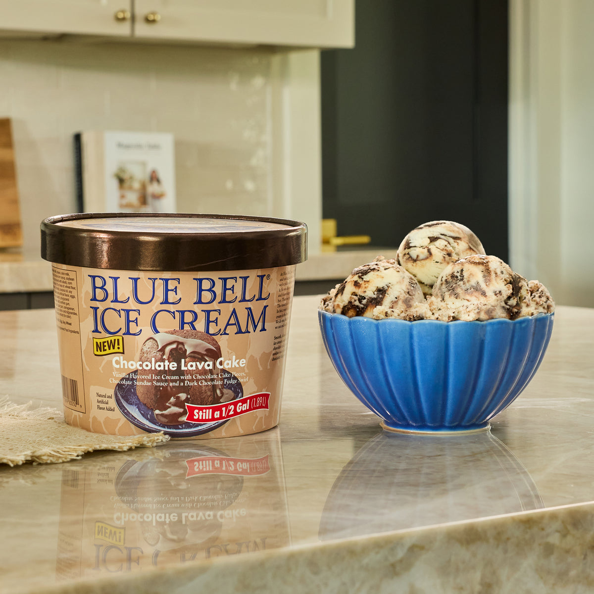 A tub of Blue Bell Chocolate Lava Ice Cream next to a blue bowl filled with chocolate lava cake ice cream on a kitchen counter.