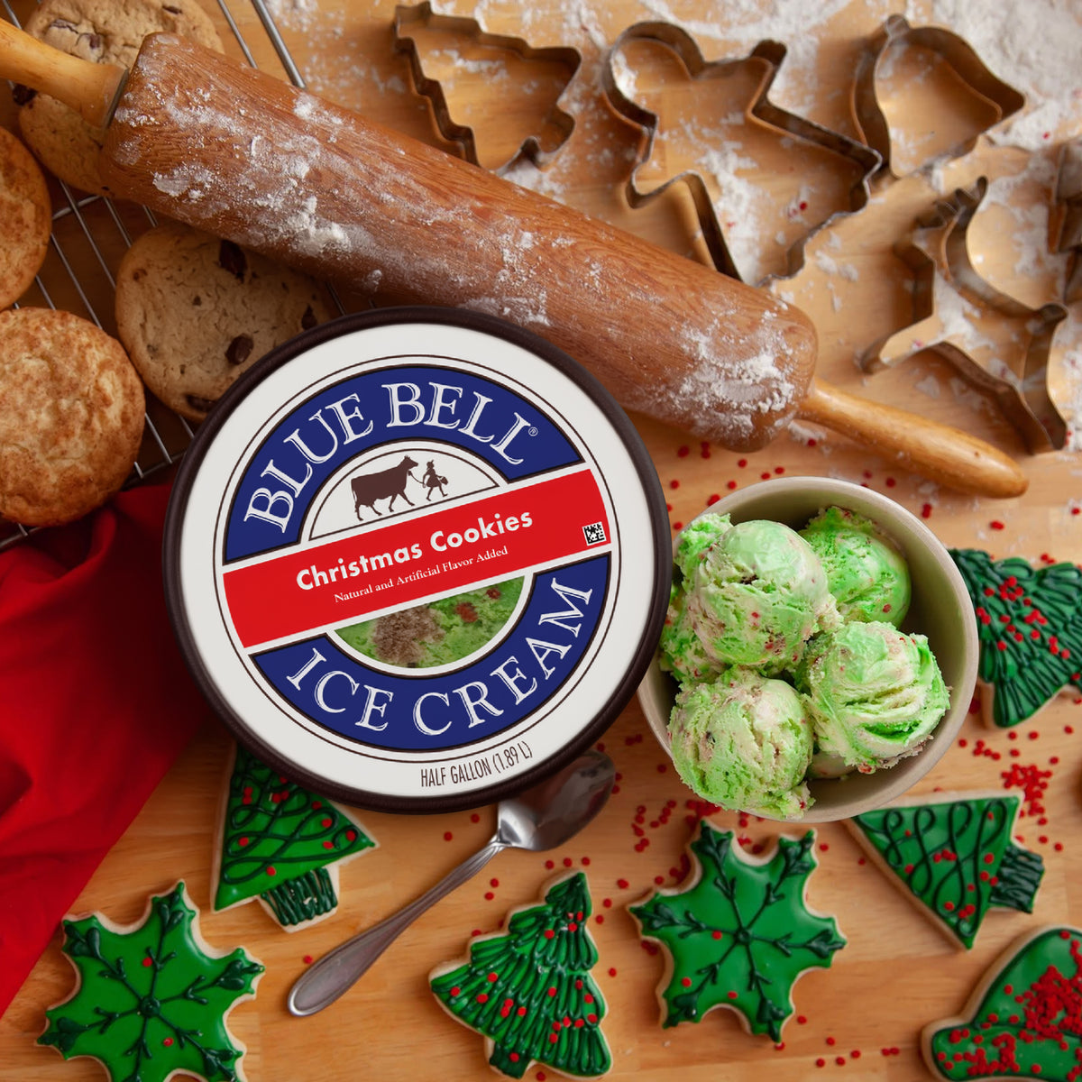 A tub of Blue Bell Christmas Cookies ice cream sits on a wooden surface surrounded by holiday baking props, including cookie cutters, a rolling pin, cookies, a bowl of green ice cream scoops, and decorated Christmas tree cookies.