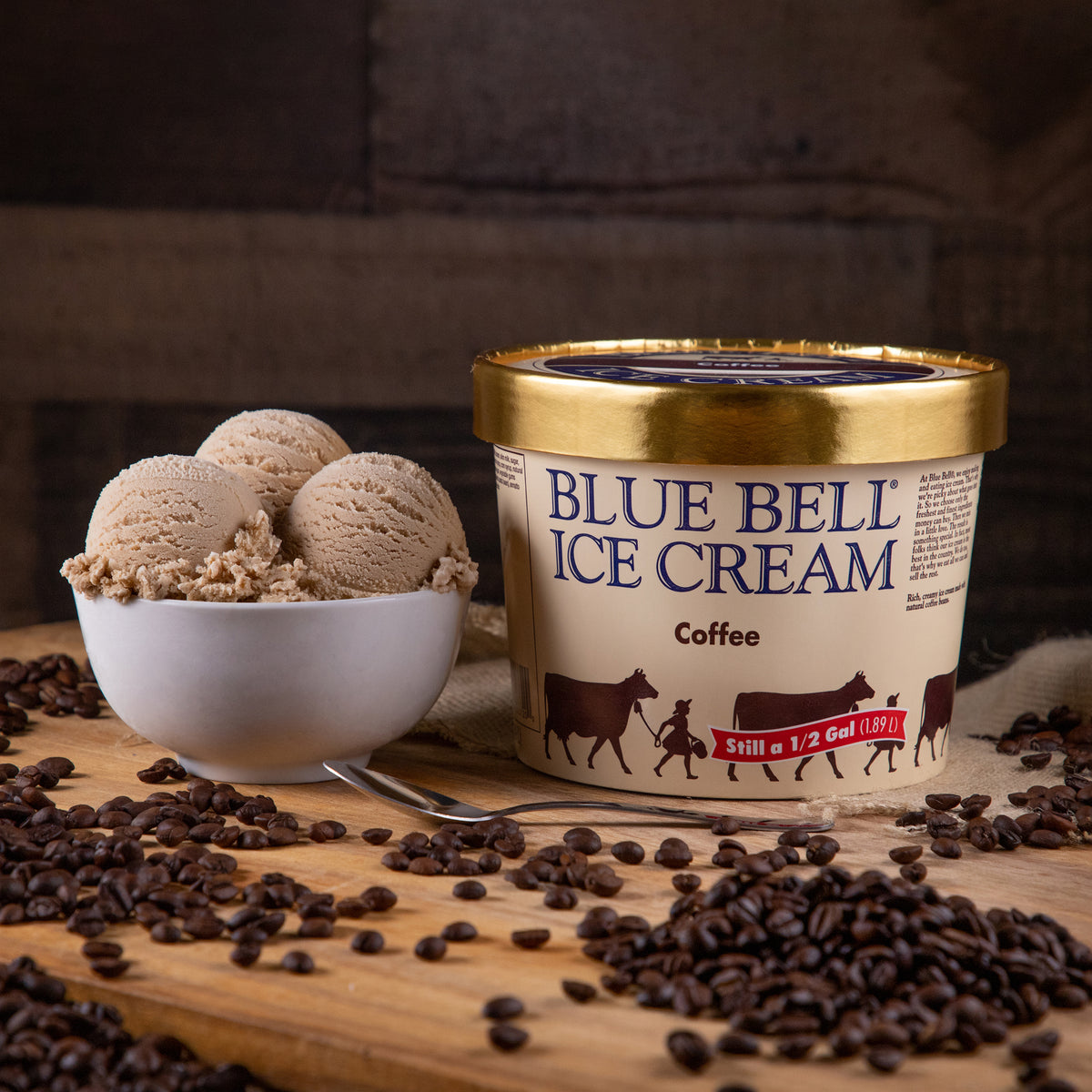 A tub of Blue Bell Ice Cream Coffee  sits on a wooden board with coffee beans scattered around it, alongside a white bowl of coffee ice cream scoops.