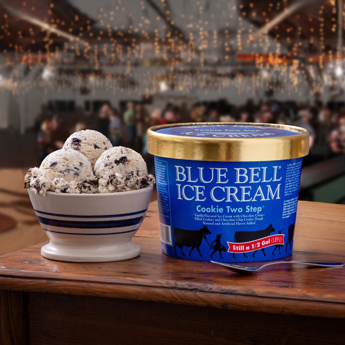 A tub of Blue Bell Ice Cream Cookie Two Step ice cream sits on a wooden table next to a bowl filled with scoops of the ice cream, with a blurred dance hall scene in the background.