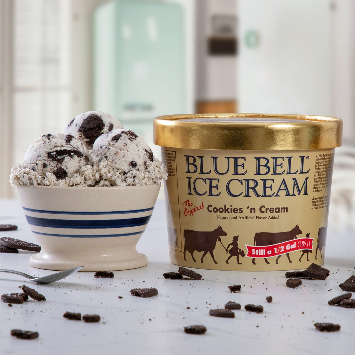 A tub of Blue Bell Ice Cream Cookies 'n Cream flavor stands next to a bowl filled with scoops of the ice cream, which has cookie crumbs and chunks on top, set on a white surface with scattered cookie pieces.