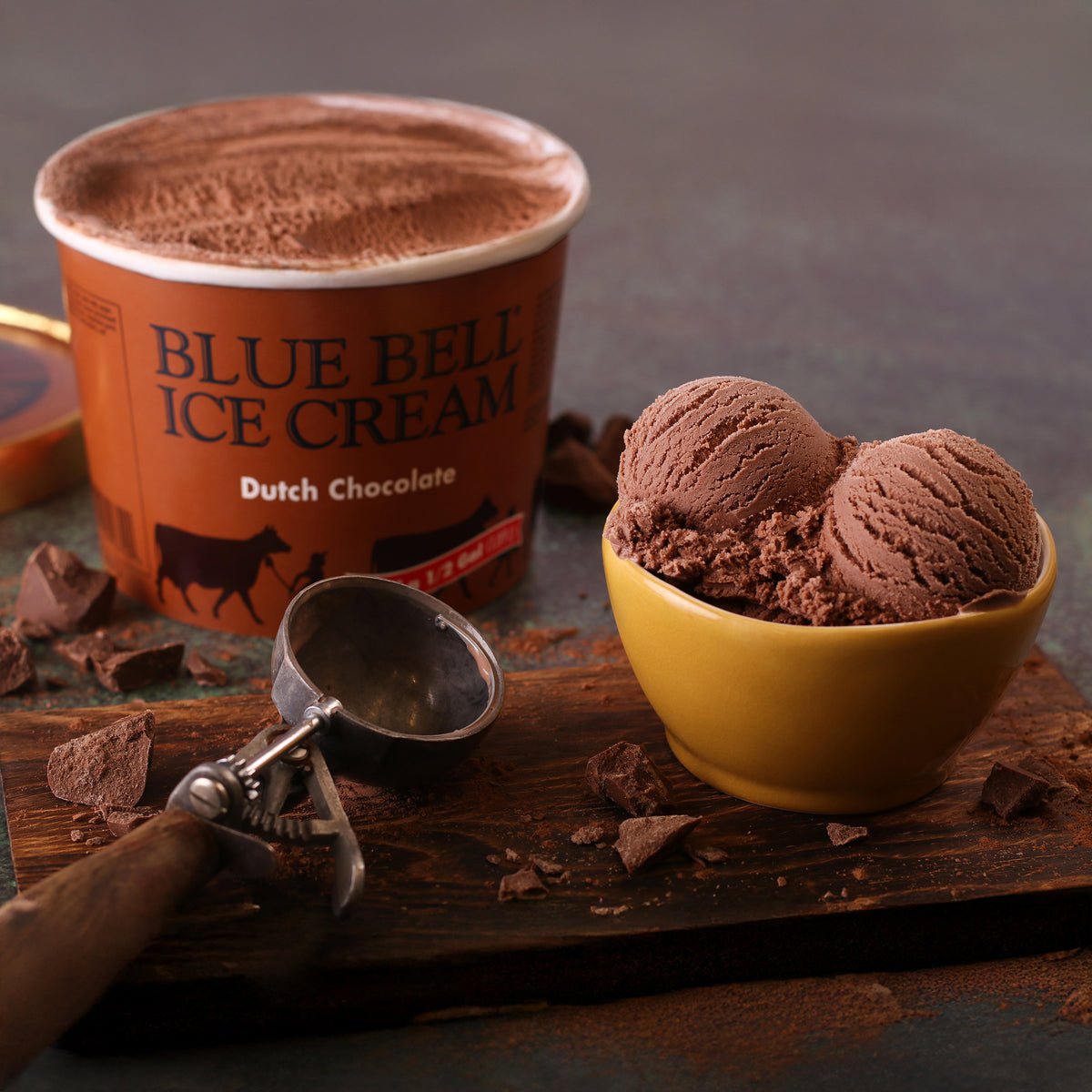 A tub of Blue Bell Ice Cream in Dutch Chocolate flavor sits on a rustic wooden board beside a yellow bowl filled with chocolate ice cream scoops, with an ice cream scoop and broken chocolate pieces nearby.