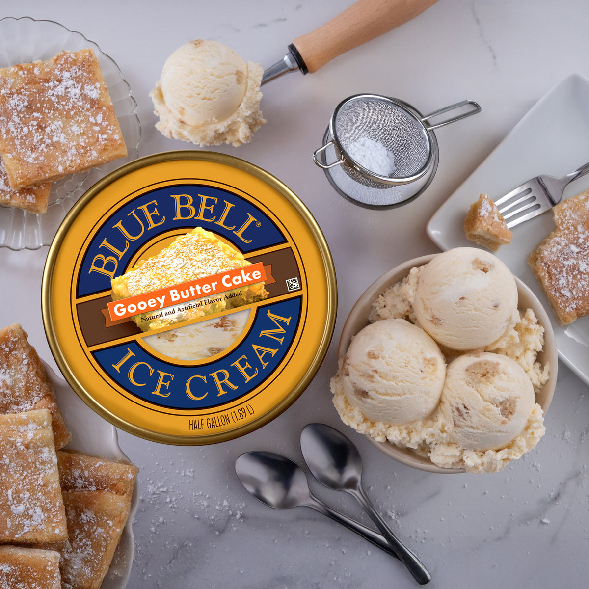 A tub of Blue Bell Gooey Butter Cake ice cream is styled in a dessert flat lay on a light marble surface, surrounded by scoops of ice cream, powdered sugar, cake pieces, spoons, and a small sifter.