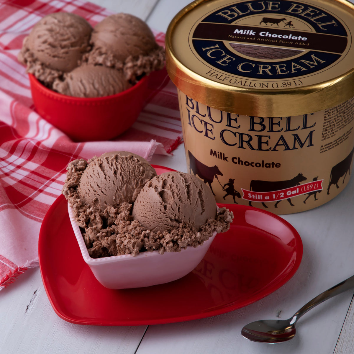 A tub of Blue Bell Milk Chocolate Ice Cream with a bowl of ice cream in a heart shaped bowl on a red heart shaped plate with a spoon on a white kitchen table.