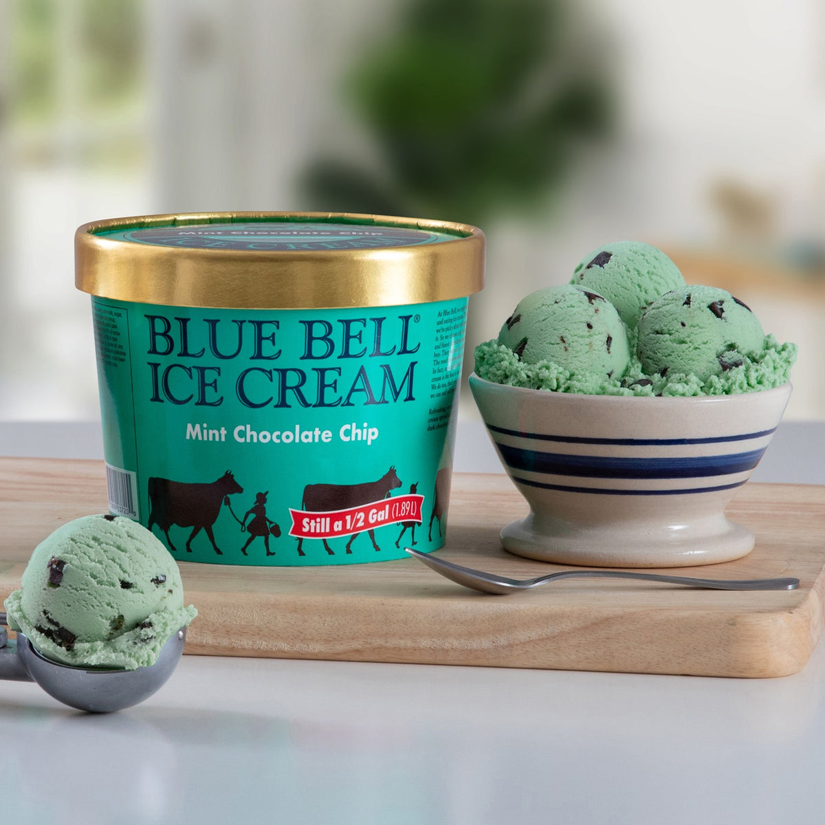 A tub of Blue Bell Ice Cream Mint Chocolate Chip is shown on a wooden board with a scoop of mint chocolate chip ice cream in the foreground and a bowl of the same ice cream in the background.