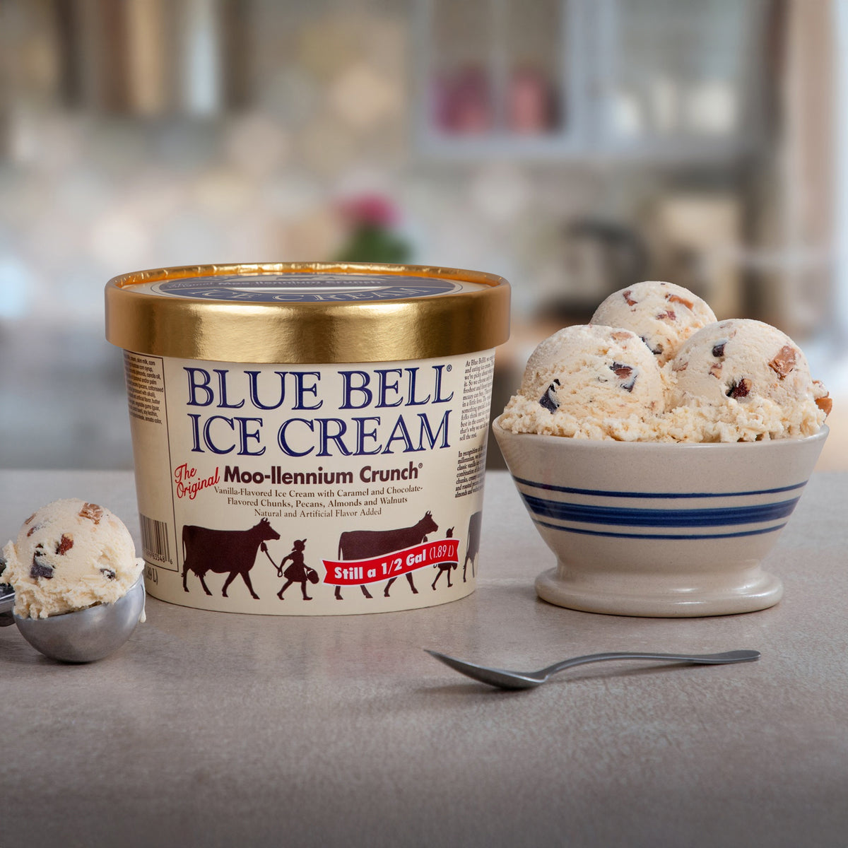 A tub of Blue Bell Moo-llennium Crunch nice cream next to a dip and bowl of ice cream with a spoon resting on a marble countertop in a blurred kitchen scene. 