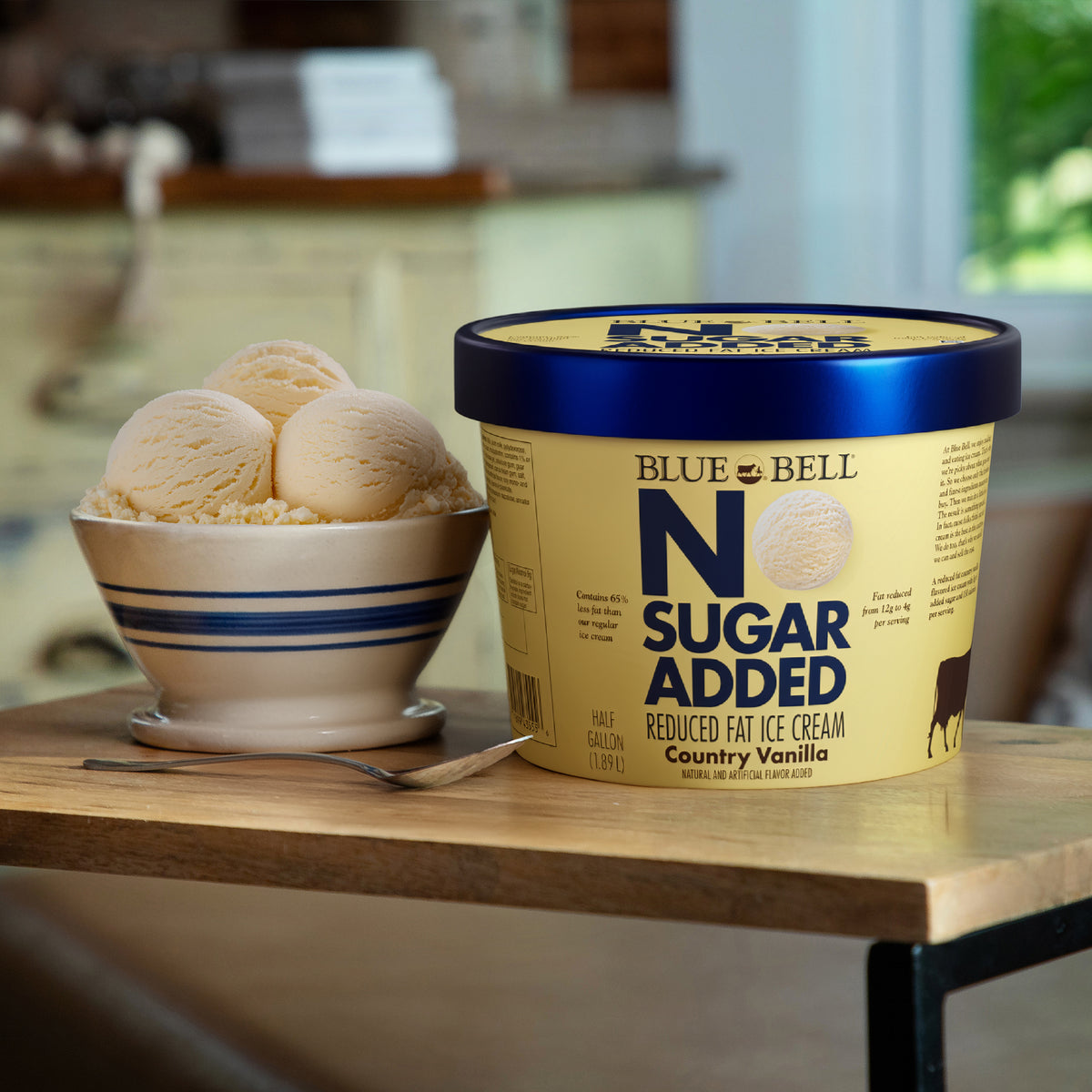 A tub of Blue Bell Blue Bell No Sugar Added reduced fat ice cream next to a bowl of vanilla ice cream scoops on a wooden table.