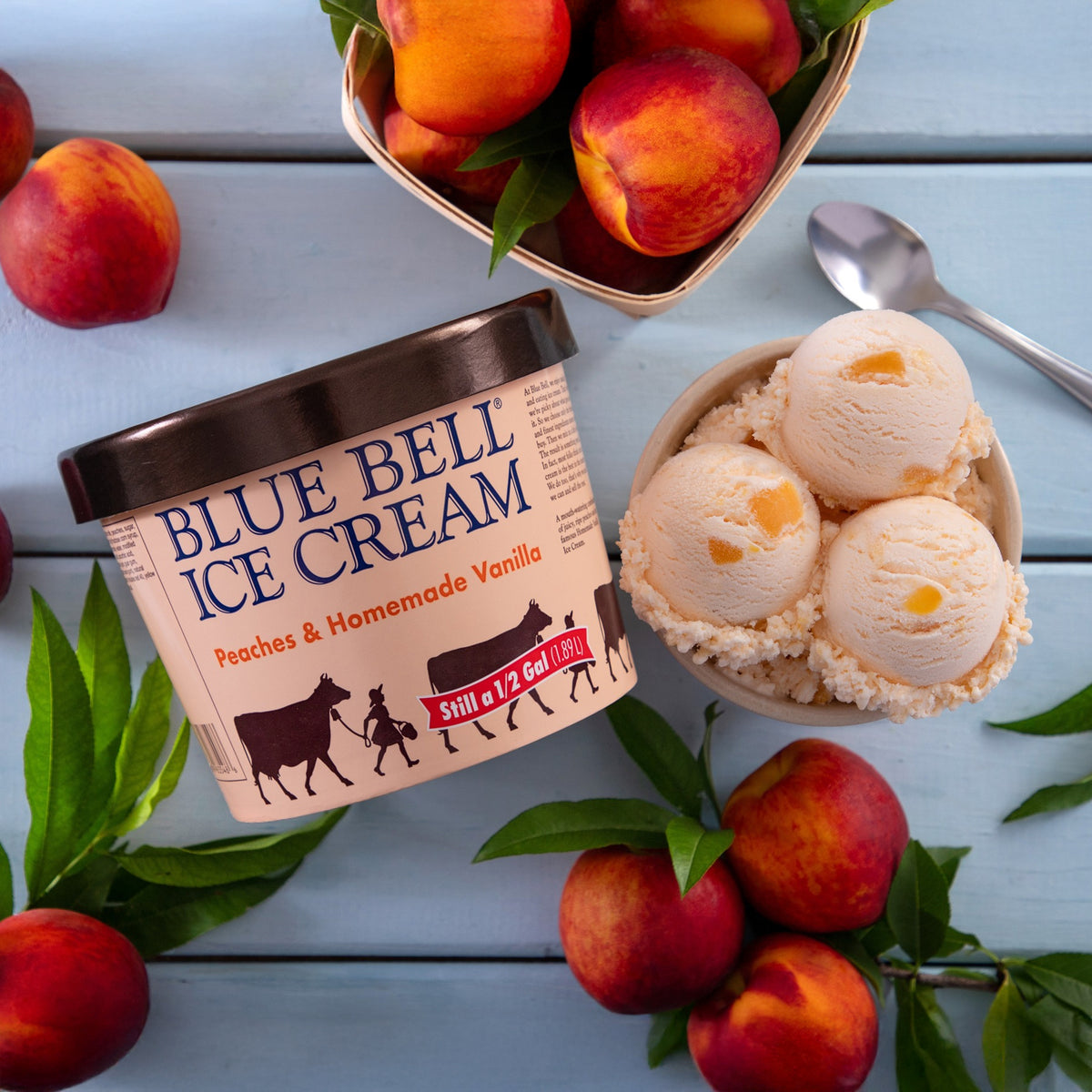 A tub of Blue Bell Ice Cream in Peaches & Homemade Vanilla flavor is styled on a light blue wooden surface with fresh peaches, green leaves, a bowl of scoops of ice cream, and a spoon.