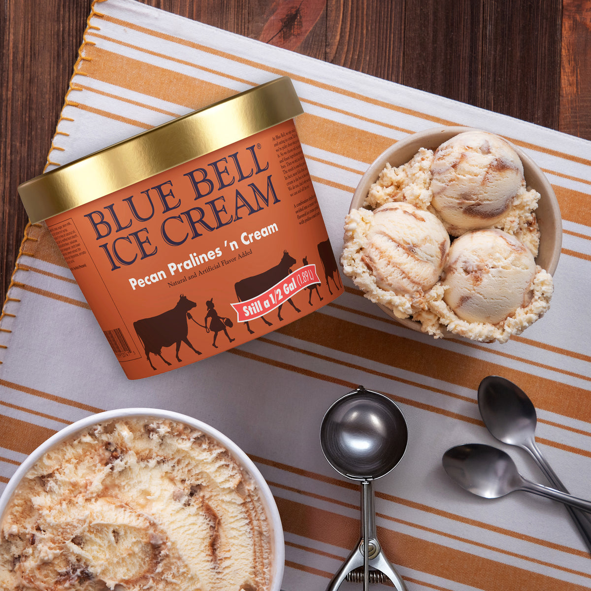 A tub of Blue Bell Ice Cream Pecan Pralines 'n Cream is shown on a striped cloth beside bowls of the ice cream and metal ice cream scoops, with a wooden table background.