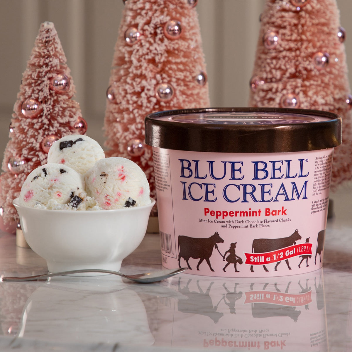 A tub of Blue Bell Ice Cream in Peppermint Bark flavor sits on a reflective surface next to a white bowl with three scoops of the ice cream, with decorative pink Christmas trees in the background.
