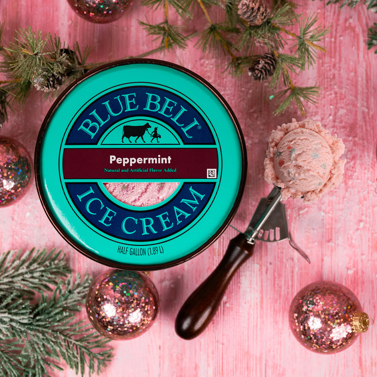A round lid of Blue Bell Ice Cream in Peppermint flavor, placed on a pink wooden surface with pine branches, glitter ornaments, and a scoop of pink peppermint ice cream beside it.