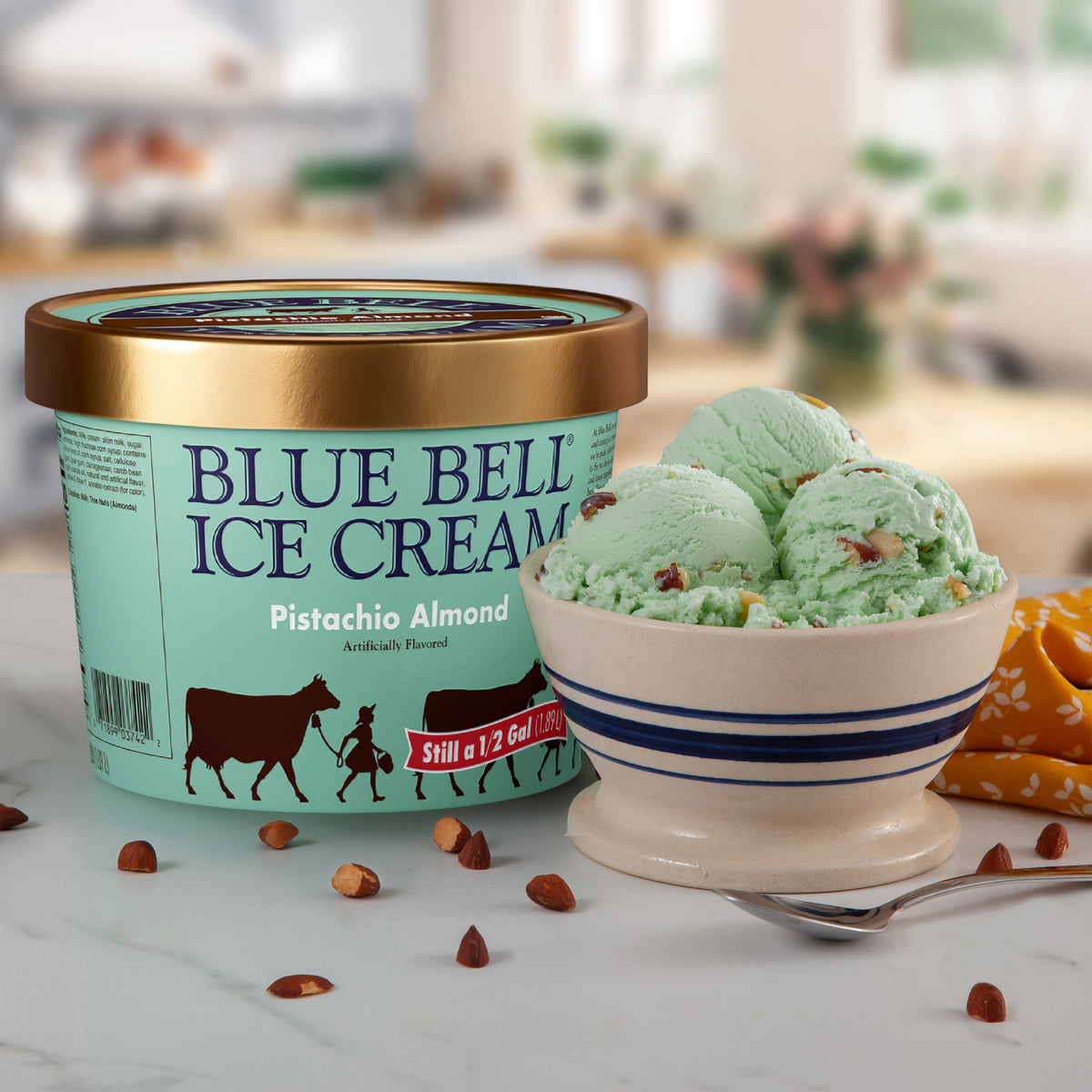 A tub of Blue Bell Ice Cream Pistachio Almond flavor sits on a marble surface next to a bowl of pistachio green ice cream scoops with nuts and a spoon, in a softly blurred kitchen setting.