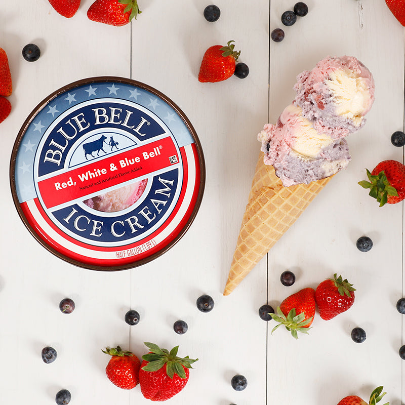 A round lid of Blue Bell Red White and Blue Bell Ice Cream on a white wooden surface surrounded by strawberries, blueberries and an ice cream cone with scoops of ice cream.